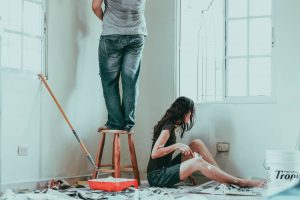 Two people painting a room. A man is standing on a stool, his upper body out of the shot. A woman is sitting on a tarp covered floor, painting around a light socket.